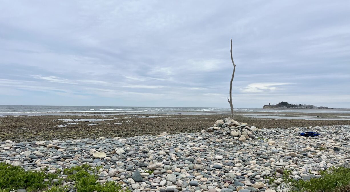 A large flat area covered with beach stones with a long stick propped up in the middle by mounded stone, and the ocean in the background.