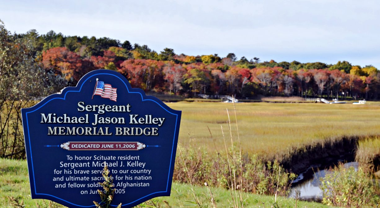 A photograph of a memorial sign with a river and marsh in the background.