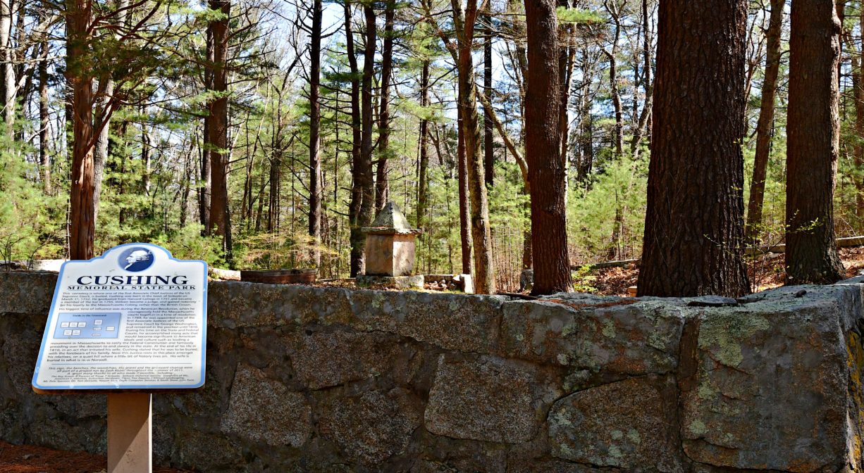 An interpretive sign beside a stone wall with a memorial in the background, in a wooded setting.