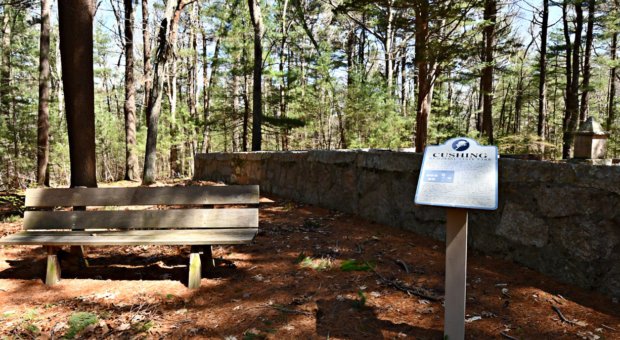A photograph of an interpretive sign and a bench with a stone wall in the background, in a forested setting.