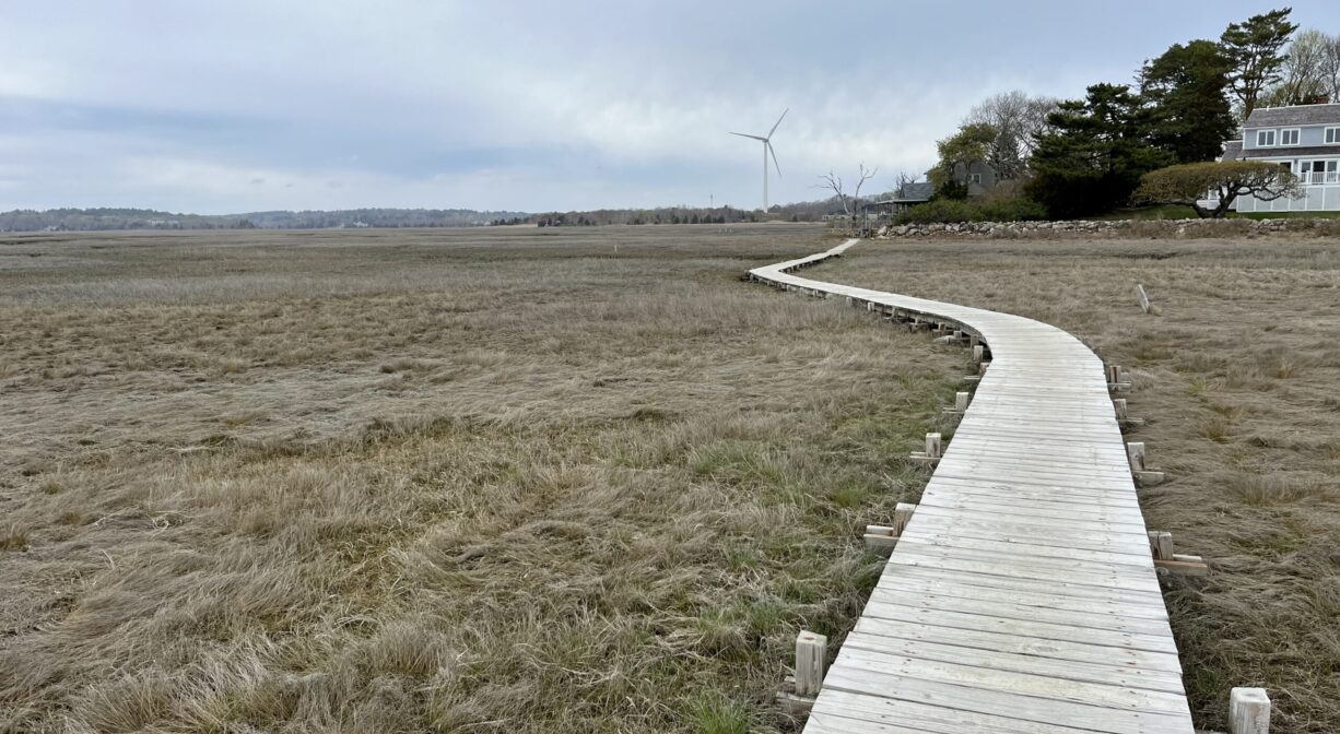 A photograph of a boardwalk arcing across a salt marsh with some houses in the distance.