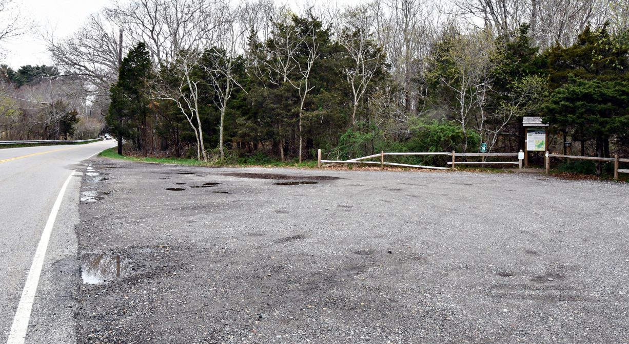 A photograph of an unpaved parking area, with some trees and signage in the background.