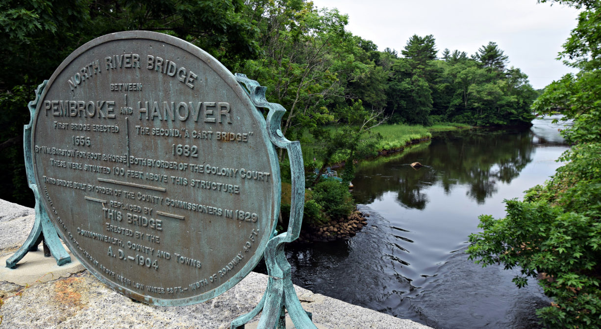 A photograph of a historic marker on a bridge overlooking a river.