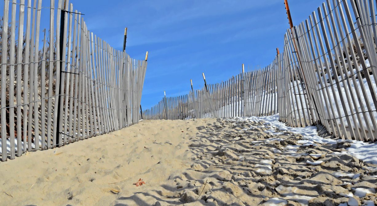 A photograph of a trail over a sand dune, with snow fence.