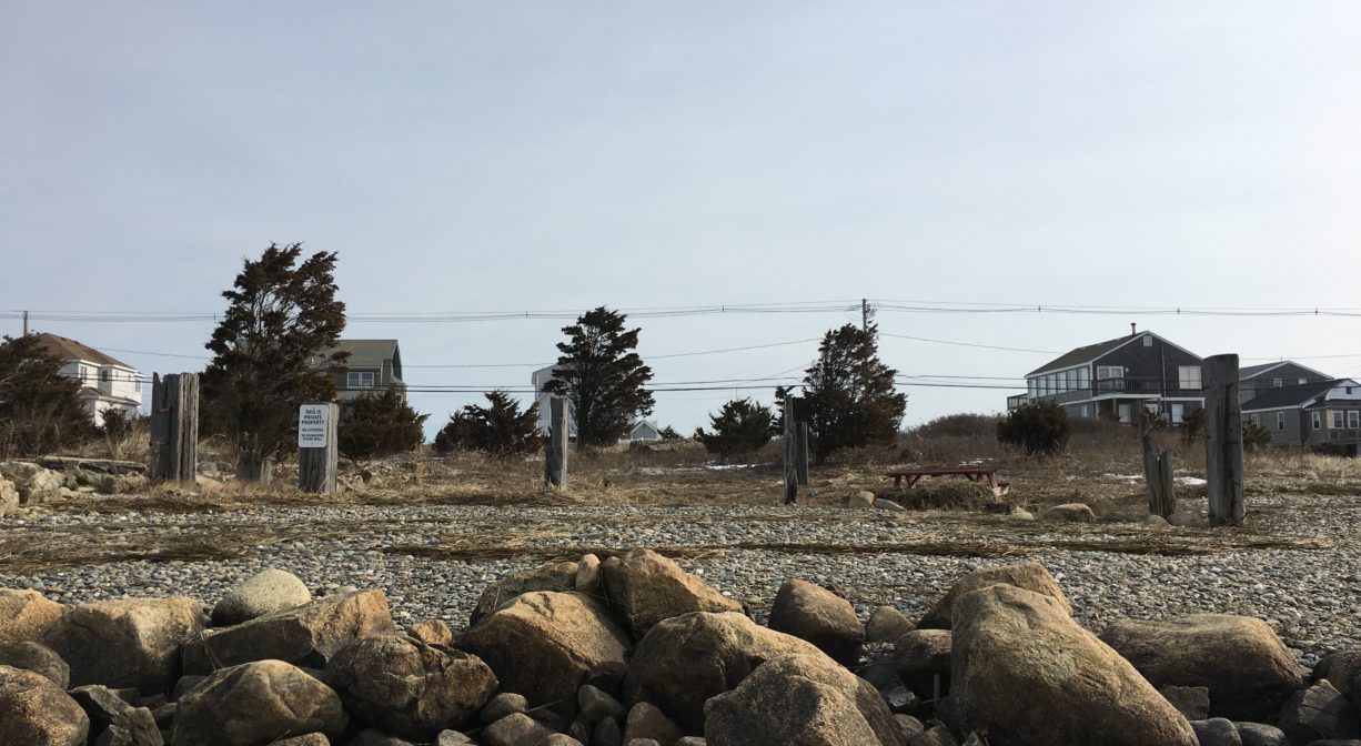 A photograph with a stone wall (or foundation) in the foreground, and a stony beachfront area in the background, with cedar posts standing up in the sand and some cedar trees in the background.