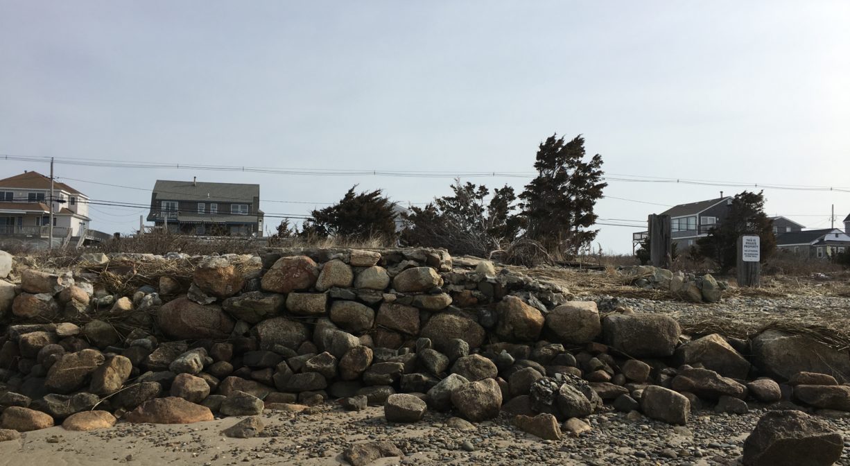 A photograph of a stone wall (or foundation) on a beach with some houses and trees in the background.
