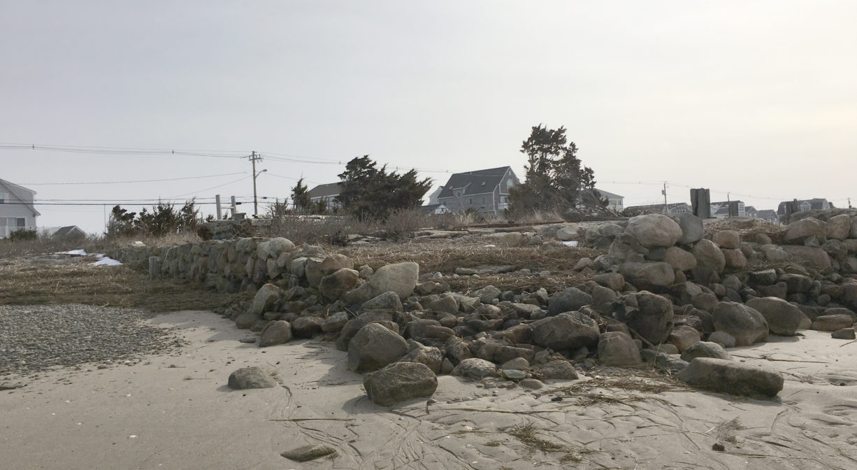 A photograph of a corner view of a stone foundation or wall, on the beach with some houses and trees in the background.