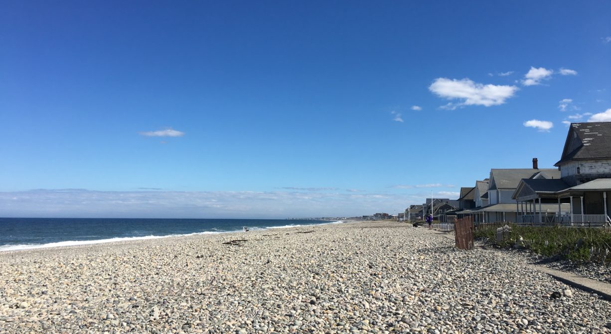 A beach covered with stones with the ocean on one side and some houses and a sea wall on the other.