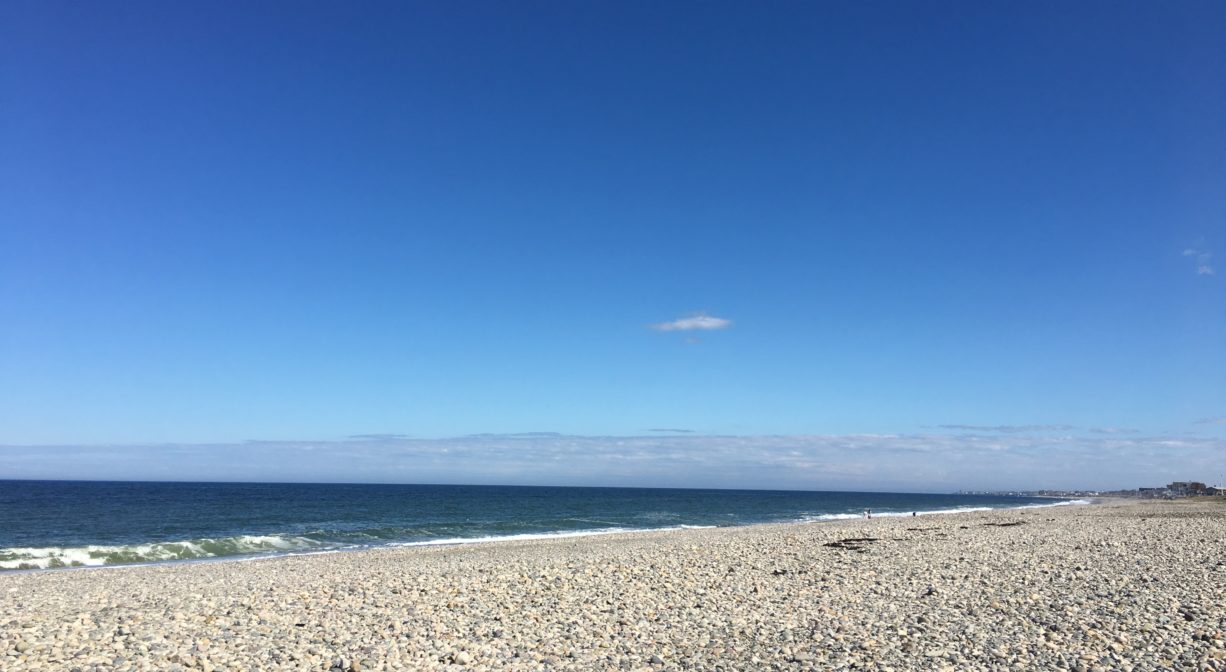 A beach covered with stones, with the ocean in the background on a sunny day.