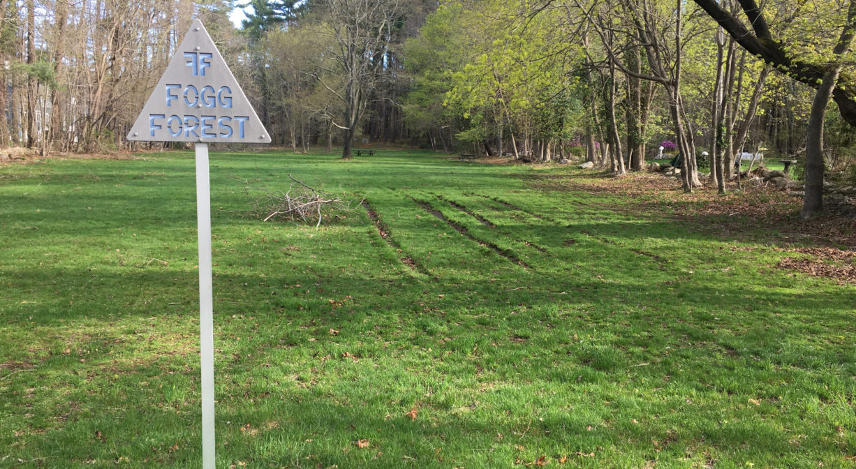 A photograph of a property sign at the edge of a field, with forest in the background.