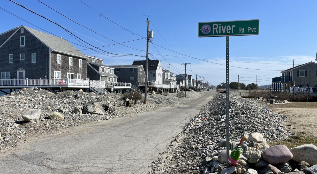 A photograph of a roadway with a street sign. The road is lined on both sides with stones, and there are houses in the distance.