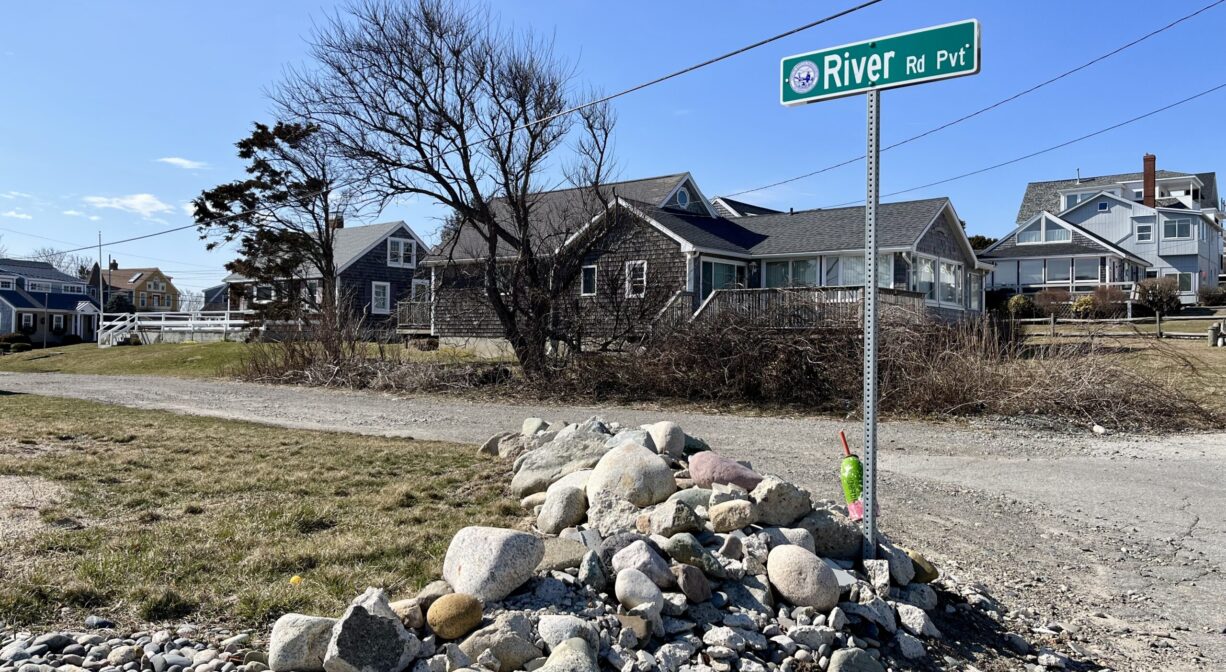 A photograph of a roadway with a street sign. The road is lined with stones, and there are houses in the distance.
