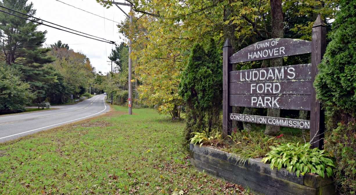 A photograph of a wooden property sign on a green lawn with trees in the background.