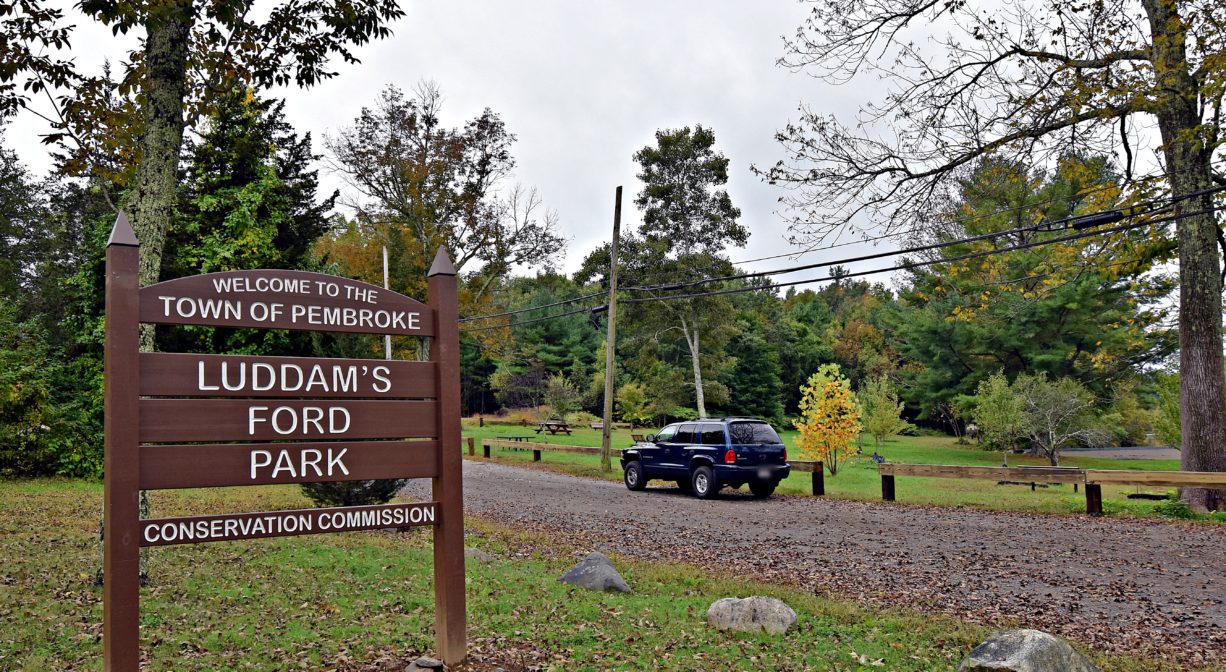 A photograph of a property sign beside a parking area, with grass and trees.