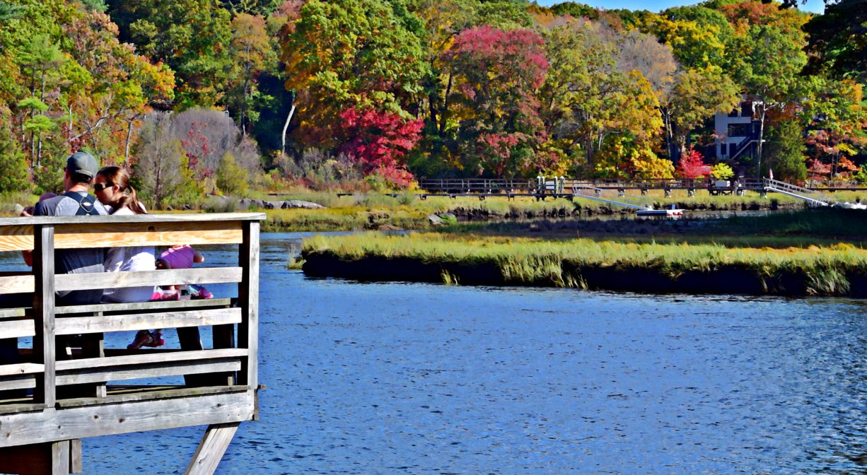 A photograph of two people standing on a deck overlooking a river, with colorful foliage.