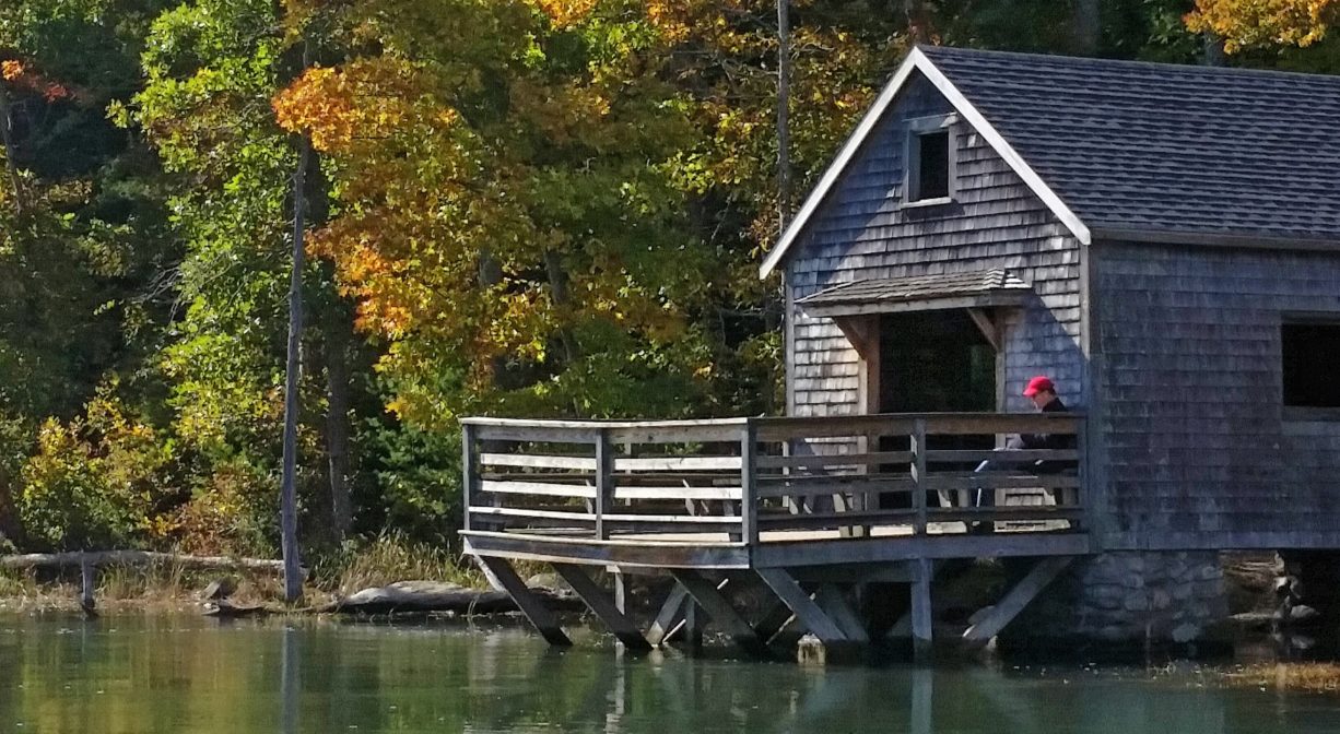 A photograph of a boat house on a river with colorful foliage.