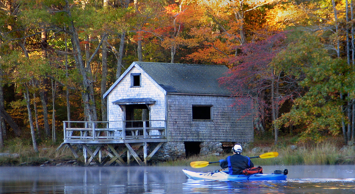 A photograph of a kayaker on a river approaching a boat house.