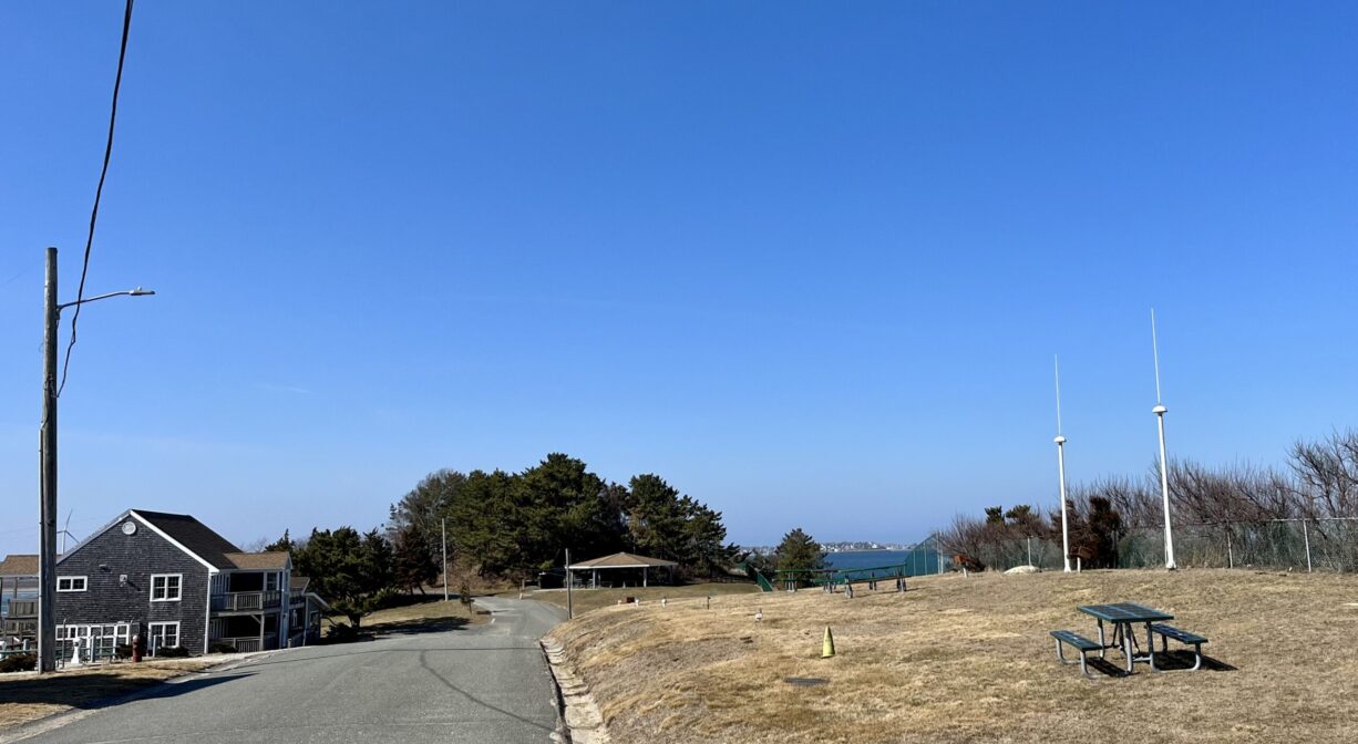 A photograph of a roadway with grass and a picnic table on one side, a house on the other and a pavilion in the distance.