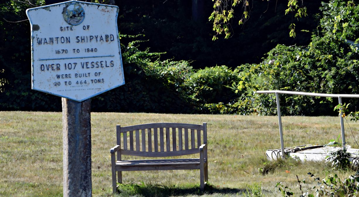 A photograph of a historic marker and a bench in a grassy area with trees in the background.