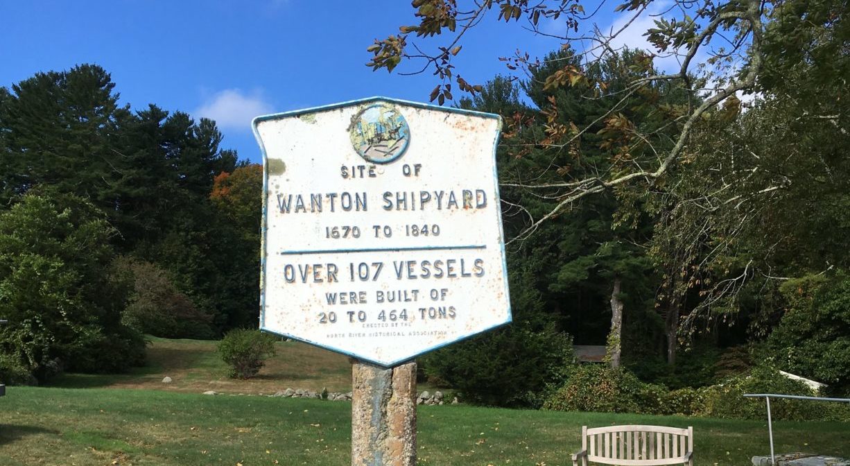 A photograph of a historic marker in a grassy area with a bench and some trees in the background.
