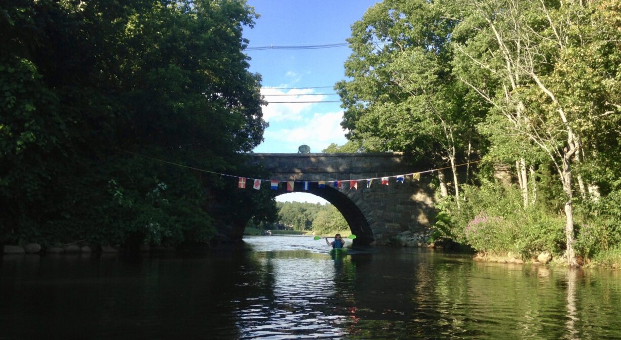 A photograph of a kayaker approaching an old stone bridge over a river, with a finish line flag above.
