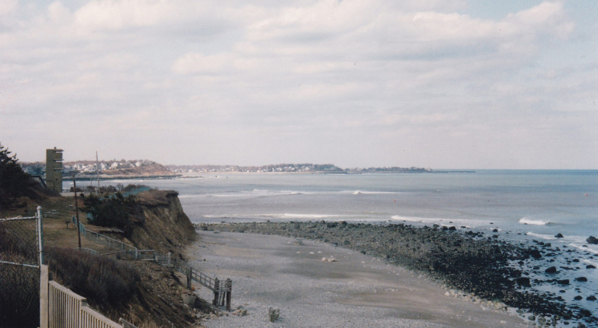 A photograph of the edge of a cliff with a beach to one side and a river and a tower in the distanc