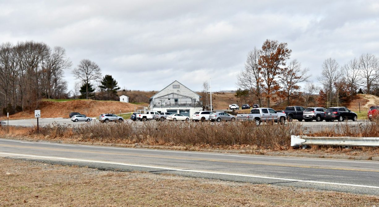 A photograph of a golf course with a clubhouse and a parking area in the foreground.