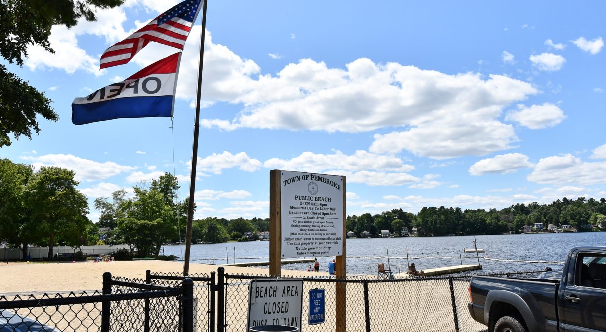 A photograph of a parking area and a flagpole at the edge of a sandy beach on a pond.