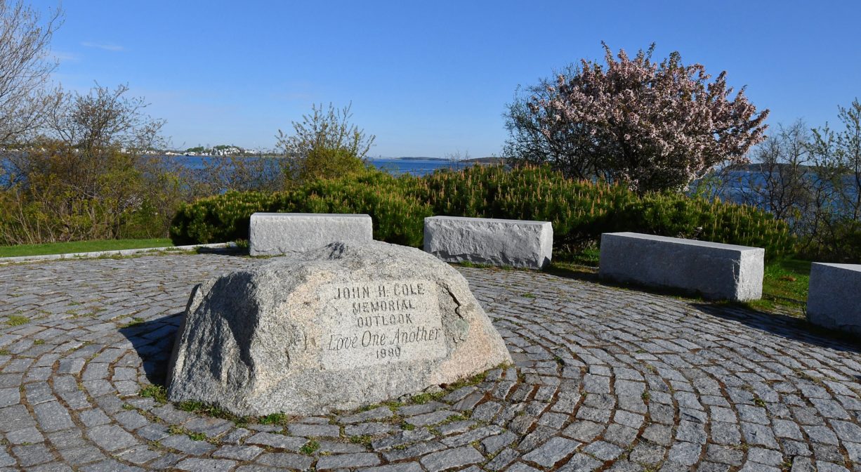 A photograph of a memorial with a boulder in the middle and pavers around it, plus benches.
