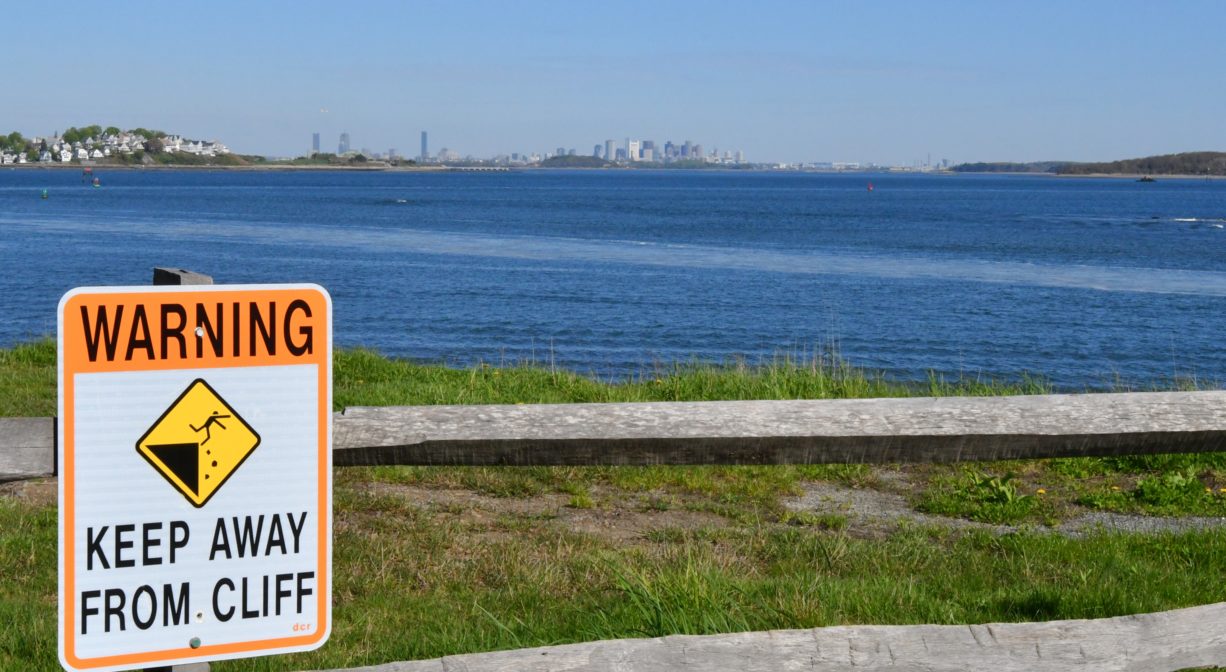 A photograph of a warning sign on a fence overlooking the water with a city skyline in the distance.