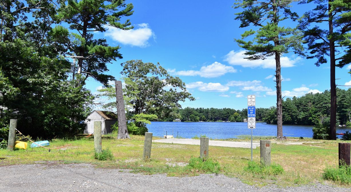A photograph of a pond with grass and trees in the foreground and a small parking area.
