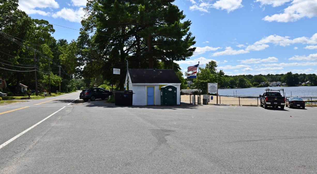 A photograph of a parking area and shed beside a sandy beach on a pond.