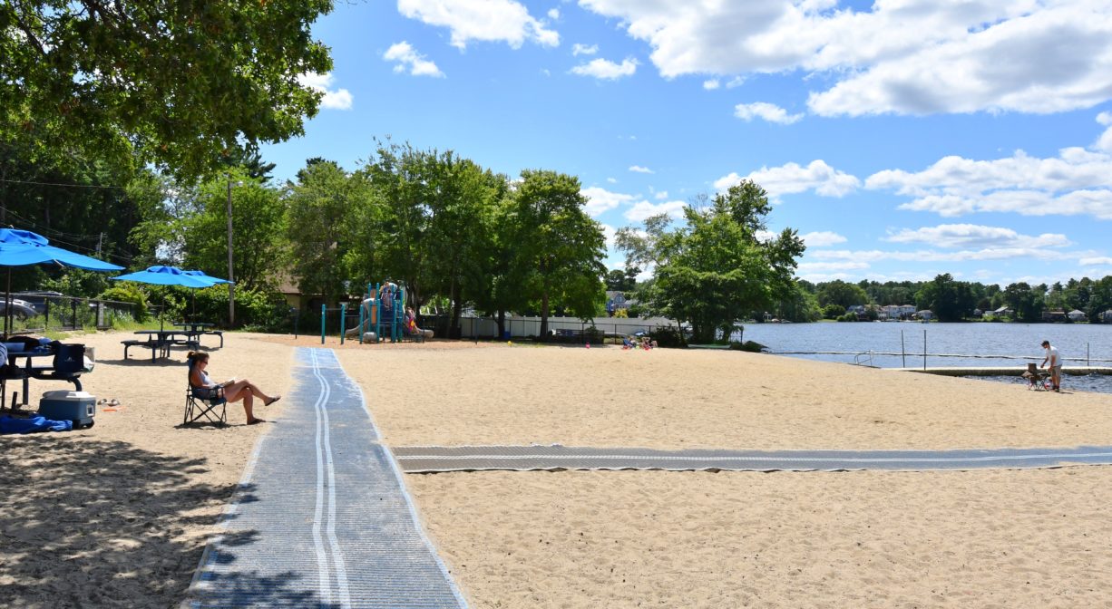 A photograph of a blue accessibility mat extending over a sandy beach beside a pond.