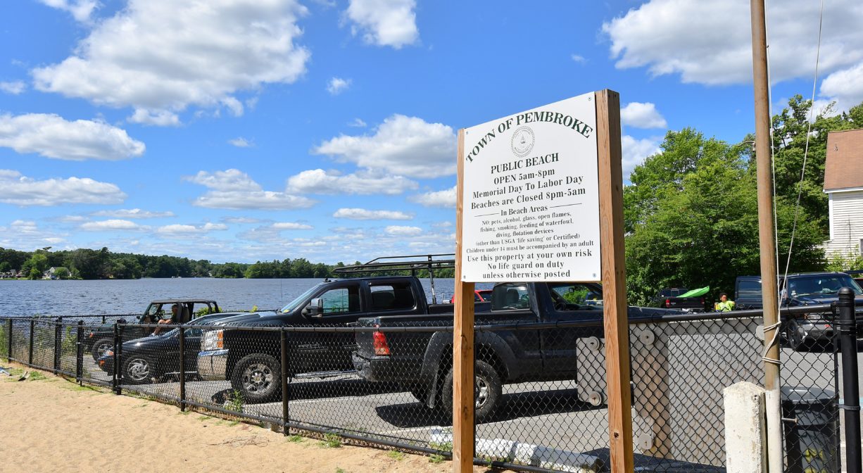 A photograph of a sandy beach bordered by fence and a parking area, with a property sign.