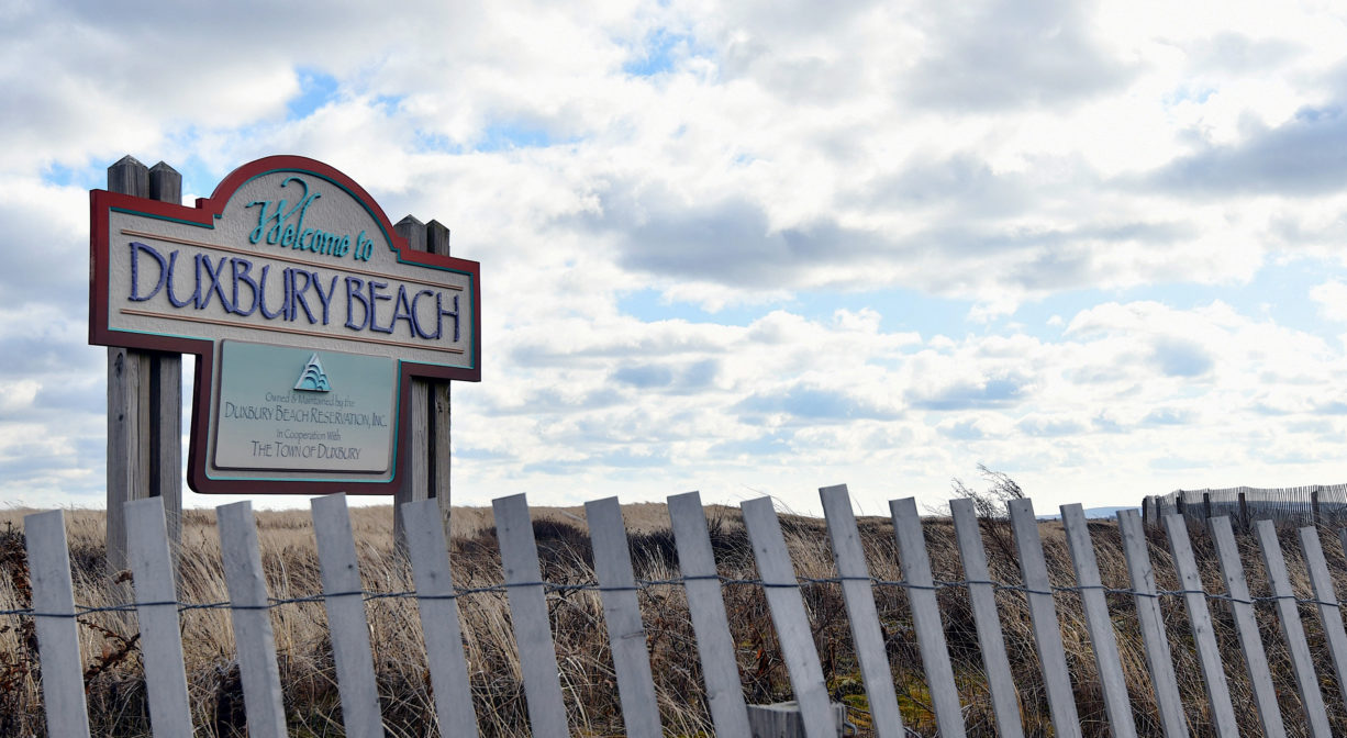 A photograph of a property sign with fence and dune grass.