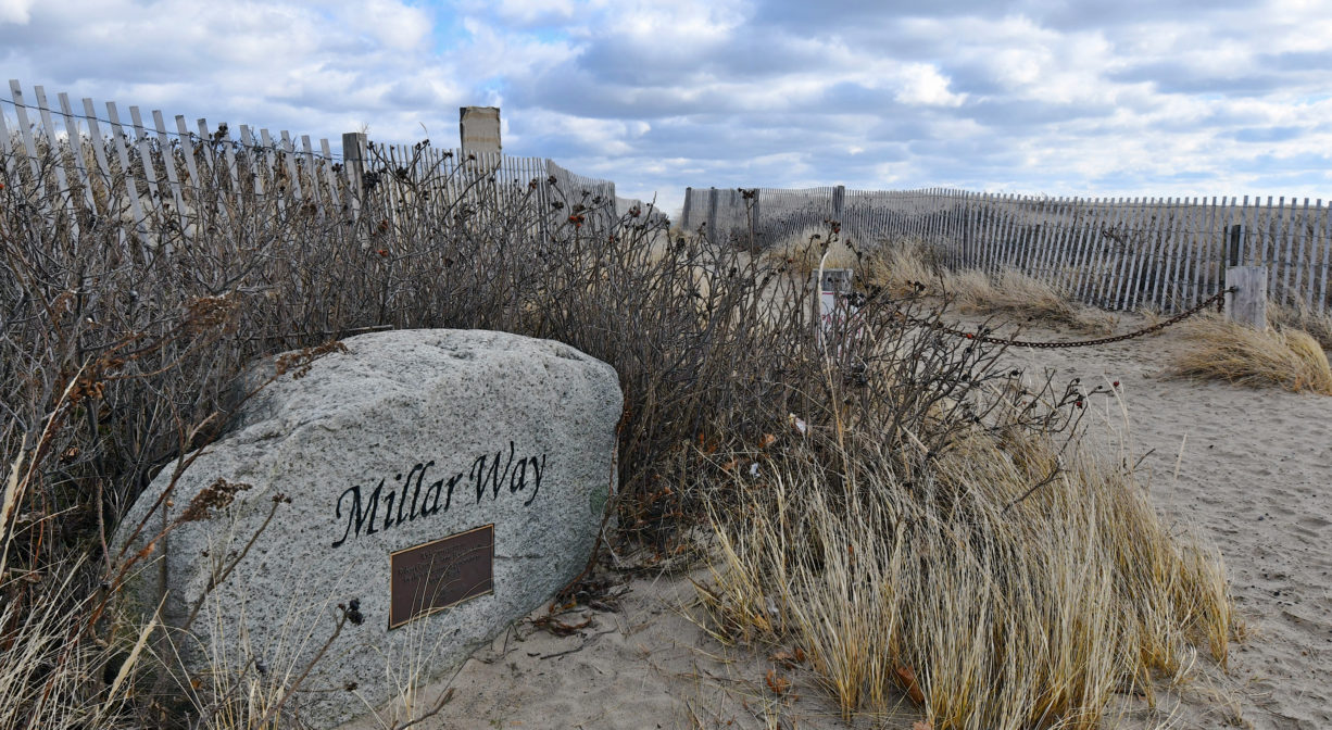 A photograph of a boulder / property sign, with dunes grass and fence.