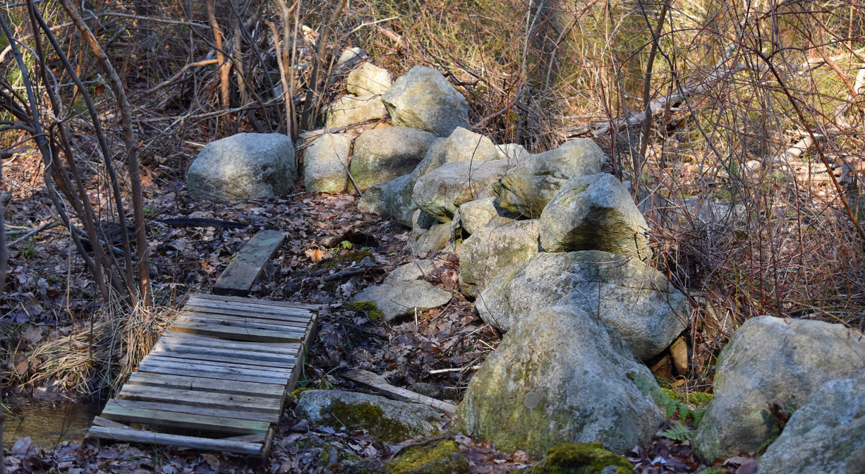 A photograph of a small wooden footbridge next to a stone wall in the woods.