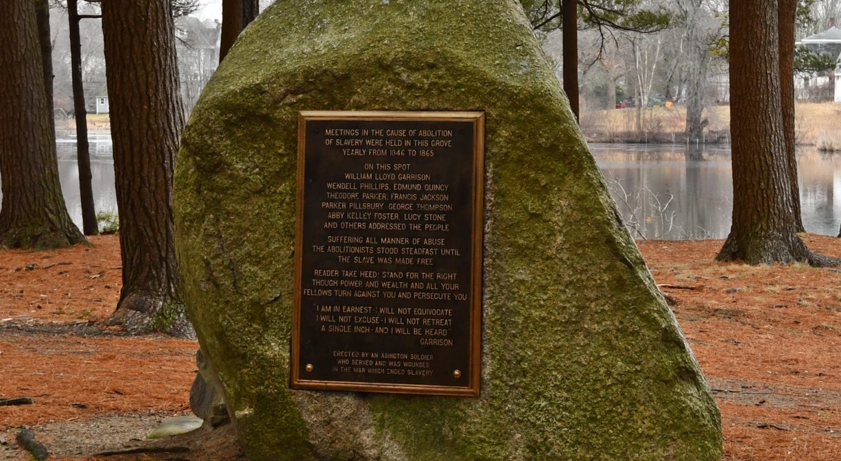 A photograph of a memorial posted on a boulder in a wooded setting.