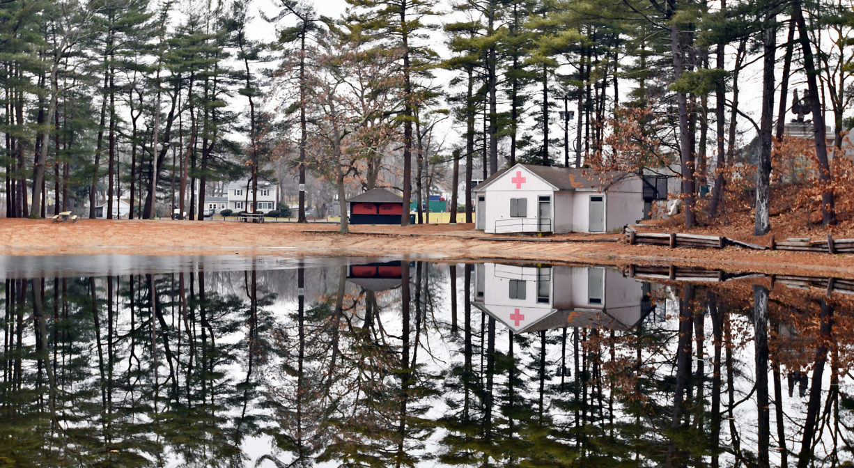 A photograph of a pond with trees reflected in its surface. There are two small buildings in the distance.