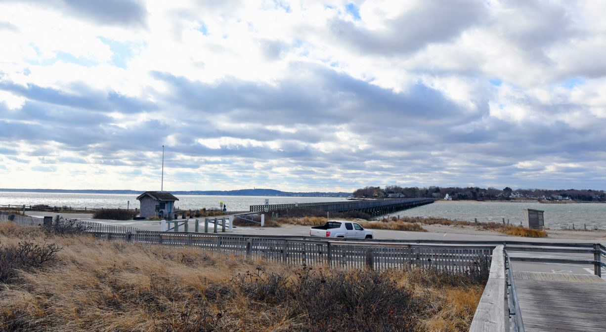 A photograph of a large wooden entrance walkway to a beach with a parking lot and a bay in the background.