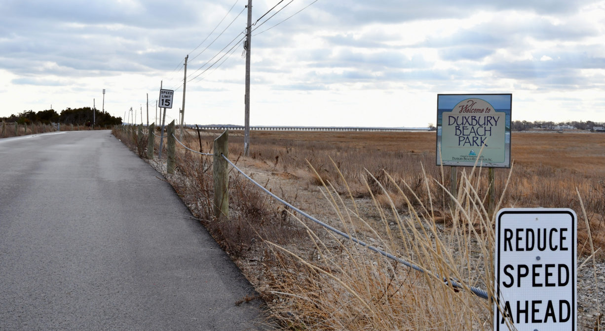 A photograph of an entrance road with property signs. A marsh and a bridge are in the background.
