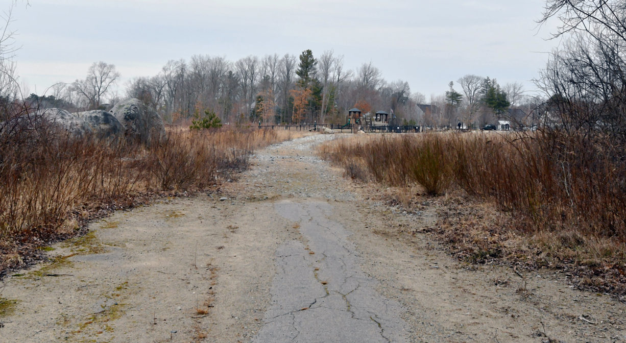 A photograph of a partially paved trail with grass and trees.