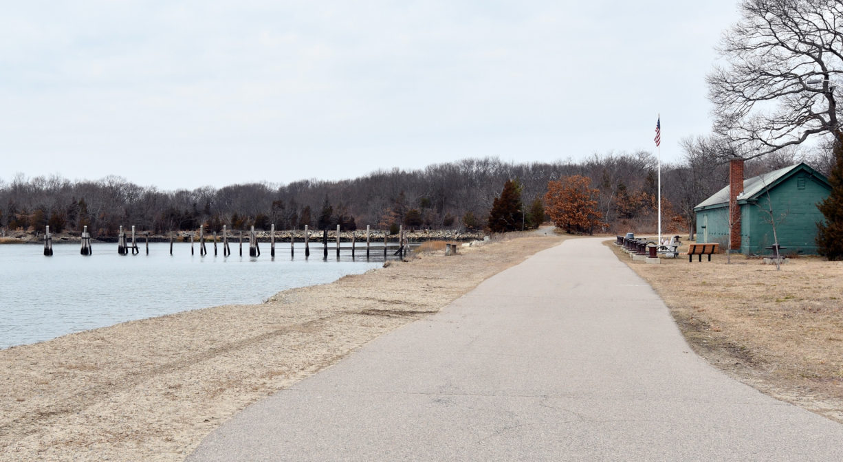 A photograph of a paved trail beside a river, with a building and a flagpole to one side.