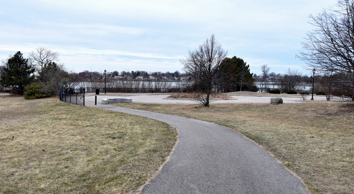 A photograph of a paved trail across a grassy area with a river in the background.