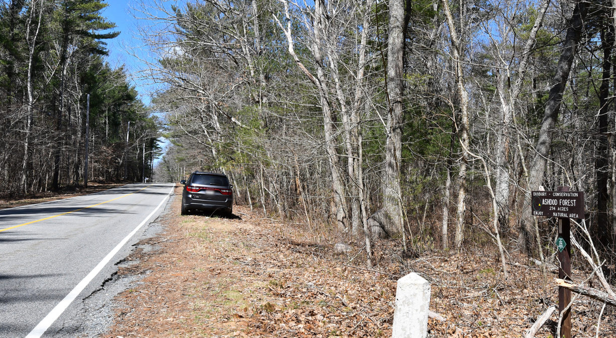 A photograph of a roadside with a car parked on the shoulder, with woods to one side.
