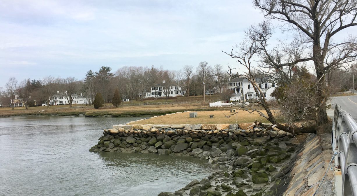 A photograph of a grassy park on a river, viewed from an adjacent bridge.