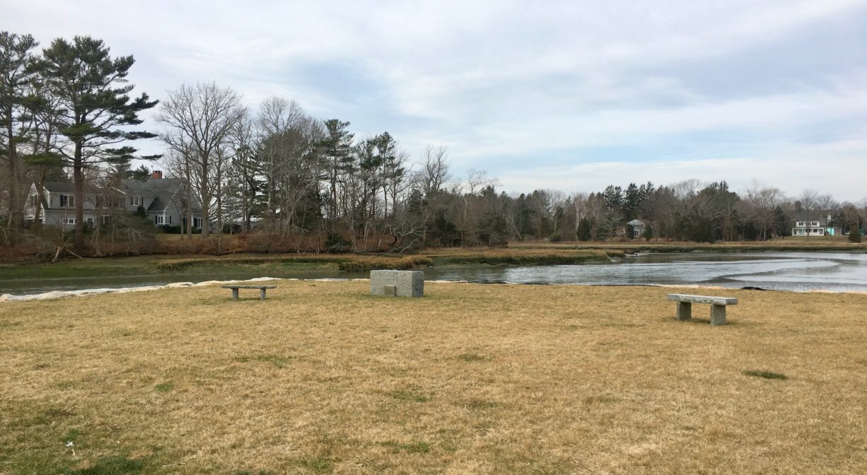 A photograph of a grassy park beside a river with granite markers and benches.