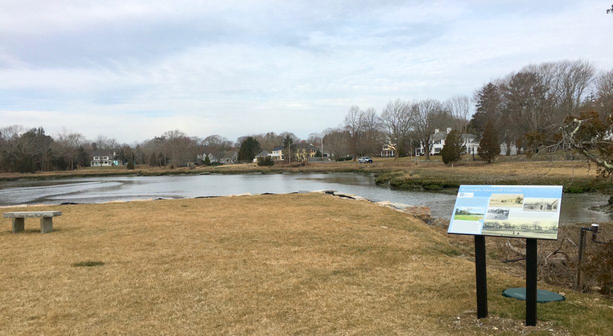 A photograph of a grassy park beside a river with a bench and an interpretive sign.
