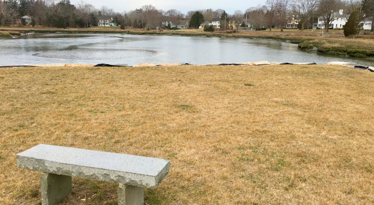 A photograph of a grassy park on a river with a granite bench.