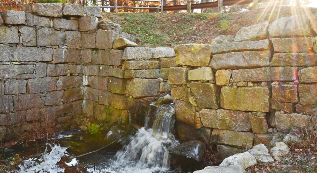 A photograph of a stream flowing through an old stone foundation.